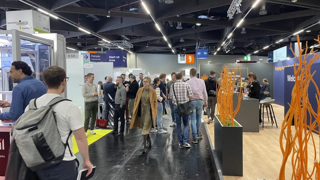 Menschen laufen durch eine belebte Kongress- oder Messehalle mit verschiedenen Ständen, Schildern und Auslagen. Einige Teilnehmer unterhalten sich, während andere sich die Exponate ansehen. Helle Lichter und modernes Dekor erfüllen den Raum.