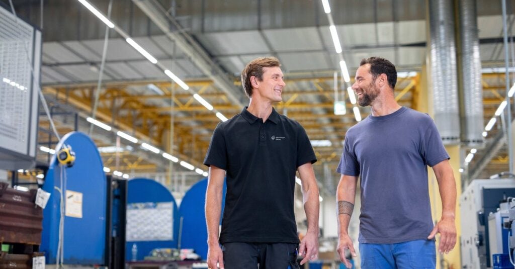Zwei Männer in legerer Arbeitskleidung gehen und lächeln sich in einer hellen, modernen Industrieanlage an, in der im Hintergrund Maschinen und gelbe Deckenbalken zu sehen sind.