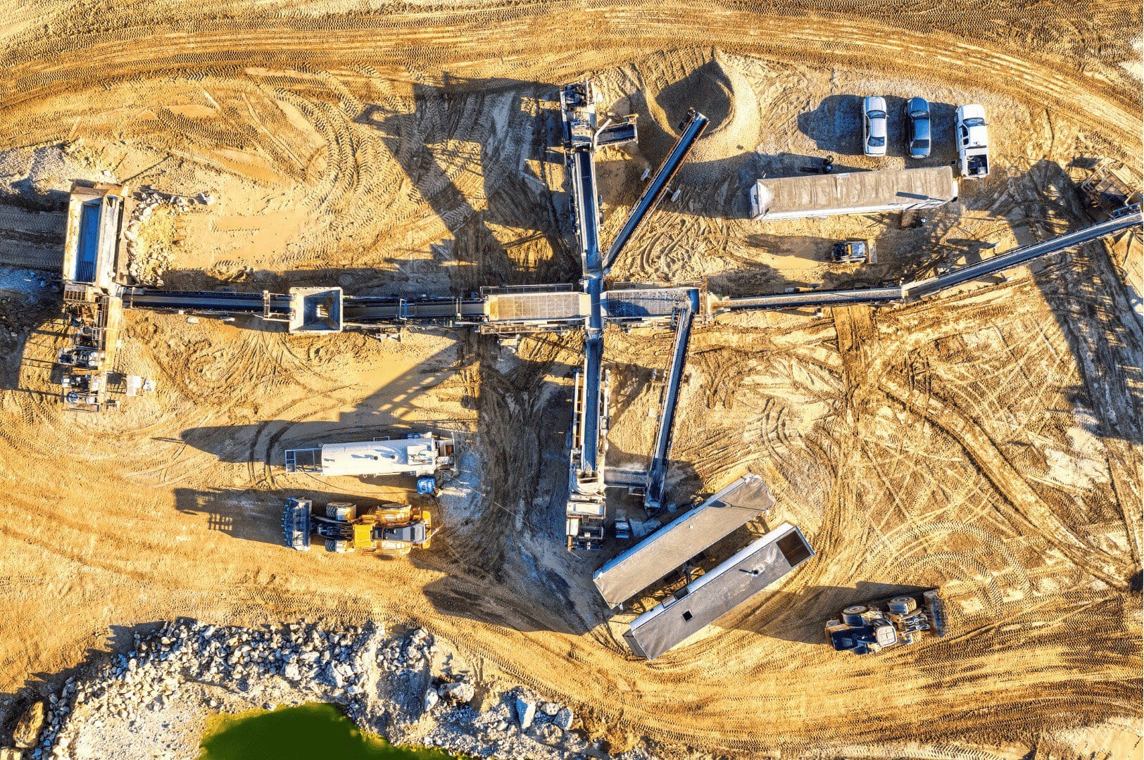 Aerial view of a construction site with heavy machinery, conveyor belts, and several vehicles on unpaved ground, surrounded by equipment and tire tracks — an example of the efficiency a Unified Namespace brings to modern mining.