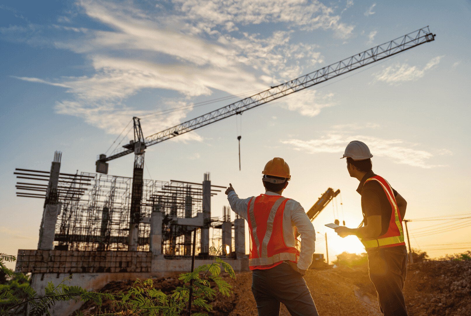 Zwei Bauarbeiter in Sicherheitswesten und mit Schutzhelmen betrachten eine Baustelle mit einem Kran und einem noch unfertigen Bauwerk, das sich vor dem Hintergrund des Sonnenuntergangs abzeichnet – eine moderne Szene aus der Baubranche, in der Konzepte wie „Unified Namespace“ (UNS) die Innovation vorantreiben.