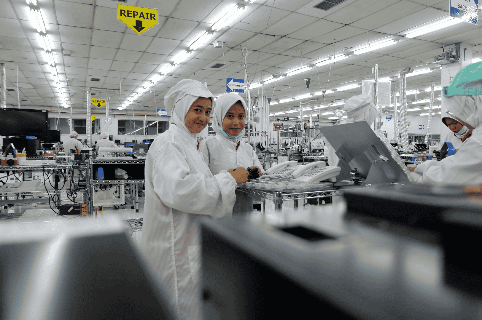 Two people in white cleanroom suits work in a bright, well-organized electronics factory, a prime example of modern electronics manufacturing. They stand near equipment and components, with a yellow 'REPAIR' sign hanging overhead, underscoring the operation's commitment to efficiency and connectivity through principles like the Unified Namespace.