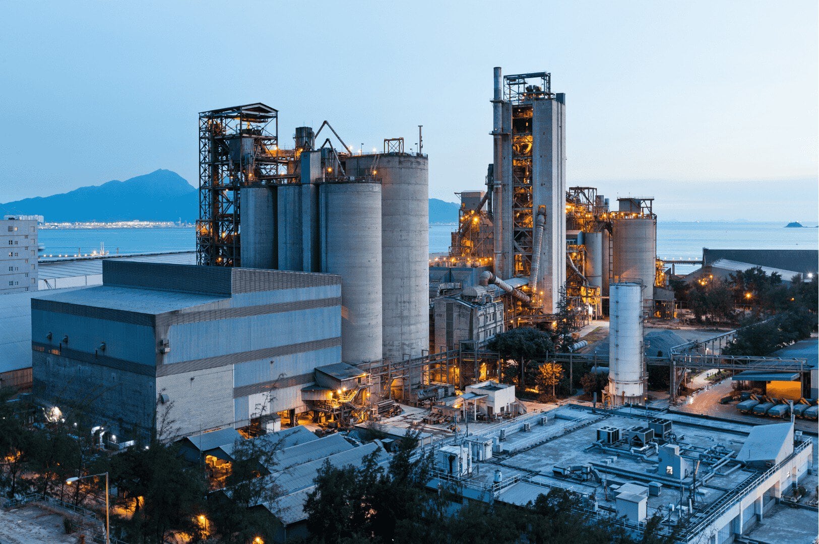 Ein industrieller Zementwerkskomplex mit hohen Silos und Türmen, der in der Abenddämmerung von künstlichem Licht beleuchtet wird und vor dem sich Berge und ein Gewässer abzeichnen, veranschaulicht moderne Abläufe in der Prozessindustrie – die möglicherweise über einen Unified Namespace (UNS) für eine nahtlose Datenintegration miteinander verbunden sind.